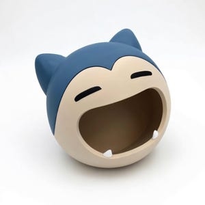 May include: A blue and tan Snorlax-shaped container with a wide open mouth and two small white teeth. The container has a smooth finish and is designed to resemble the popular Pokémon character. The ears are a darker shade of blue.