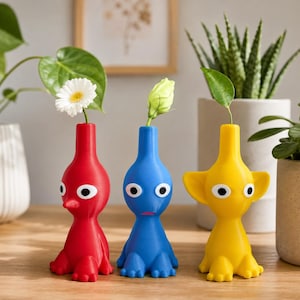 May include: Three character-shaped vases in red, blue, and yellow. Each vase features a narrow neck, a wide base with feet, and two large white eyes with black pupils. The red vase holds a white daisy, the blue vase holds a green bud, and the yellow vase holds a single green leaf.