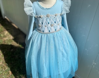 May include: A light blue tulle dress with long sleeves and a white bodice featuring a snowflake design and pearl accents. The dress has a sparkly, iridescent finish.