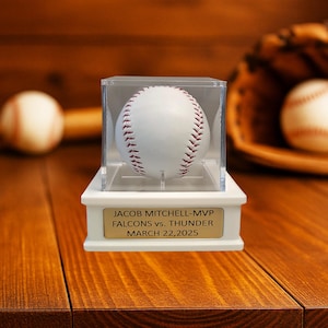 May include: A white baseball with red stitching is displayed in a clear acrylic case atop a white base. The base has a gold plaque with the text "JACOB MITCHELL-MVP FALCONS vs. THUNDER MARCH 22,2025". A baseball glove and another baseball are in the background.