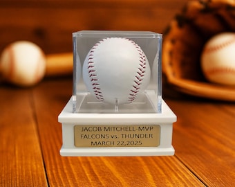 May include: A white baseball with red stitching is displayed in a clear acrylic case atop a white base. The base has a gold plaque with the text "JACOB MITCHELL-MVP FALCONS vs. THUNDER MARCH 22,2025". A baseball glove and another baseball are in the background.