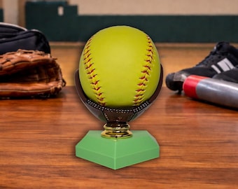 May include: A softball trophy featuring a yellow softball with red stitching, held in a brown glove-shaped holder. The base is green with a gold accent. Other sports equipment is in the background.