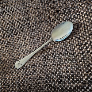 May include: A silver spoon with a rounded bowl and a long handle. The handle has the letter "M" engraved on it.