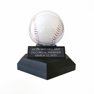 May include: A baseball in a clear display case atop a black base. The baseball has red stitching. A black plaque on the base reads "JACOB MITCHELL-MVP FALCONS vs. THUNDER MARCH 22,2025."