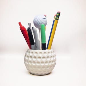 May include: A white ceramic golf ball-shaped desk organizer filled with pens, pencils, and a golf ball on a tee. The organizer has a dimpled surface, and the pens and pencils are various colors.