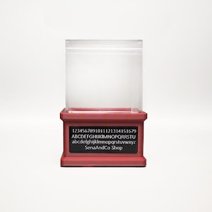 May include: A clear, rectangular display case with a red base. The base features a black rectangular label with white text, including numbers, the alphabet, and "SenaAndCo Shop."