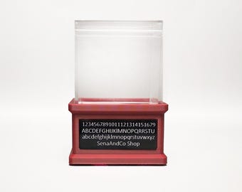 May include: A clear, rectangular display case with a red base. The base features a black rectangular label with white text, including numbers, the alphabet, and "SenaAndCo Shop."