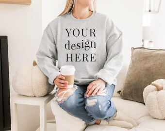 May include: A gray sweatshirt with the text "YOUR design HERE" printed on the front. The sweatshirt is being worn by a person sitting on a white couch with a white pumpkin and a gray pillow.