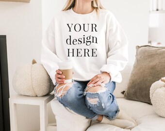 May include: A white sweatshirt with the text "YOUR design HERE" printed on the front. The sweatshirt is being worn by a person sitting on a white couch, holding a to-go coffee cup.