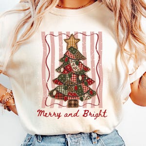 May include: An ivory t-shirt featuring a graphic of a patchwork-style Christmas tree with a star on top, set against a background of vertical pink stripes. The text Merry and Bright is printed in red below the tree design on the front of the shirt.
