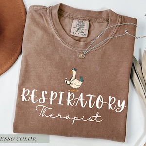 May include: Espresso-colored t-shirt with the words "Respiratory Therapist" in white script. The shirt features a cartoon duck wearing a hat and holding a medical device. A silver necklace and a brown hat are also visible.