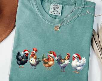 May include: A light green Comfort Colors t-shirt featuring a festive design of five chickens wearing Santa hats and string lights. A silver necklace with a pendant is draped on the shirt. The text "LIGHT GREEN COLOR" is at the bottom.