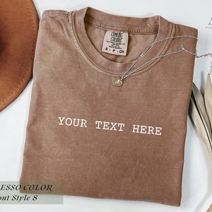May include: Espresso-colored t-shirt with the text "YOUR TEXT HERE" in white. A silver necklace with a pendant rests on the shirt. A brown hat and dried leaves are in the background. The shirt is labeled "Comfort Colors".