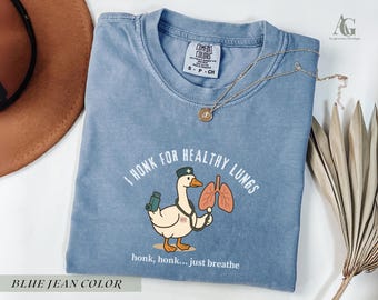 May include: Blue jean colored Comfort Colors t-shirt with a cartoon goose wearing a nurse's hat and holding lungs. Text reads "I honk for healthy lungs" and "honk, honk... just breathe". A gold necklace is draped on the shirt.
