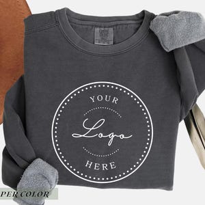 May include: A charcoal gray crewneck sweatshirt featuring a white circular graphic on the chest. The graphic contains a dotted border and the text YOUR LOGO HERE in a combination of serif and script fonts. The sweatshirt has a fleece-lined interior.
