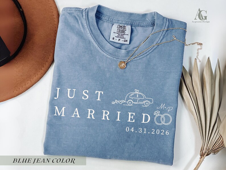 May include: A blue jean colored t-shirt with the words "JUST MARRIED" in white. A car illustration, rings, and the date 04.31.2026 are also printed on the shirt. A gold necklace is draped across the shirt.