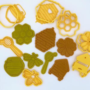 Könnte beinhalten: Eine Sammlung gelber und grüner Ausstecher in verschiedenen Formen, darunter Bienenwaben, Bienen, Honigtöpfe und Bienenstöcke. Die Ausstecher sind auf einer weißen Oberfläche angeordnet, ideal für thematische Leckereien.