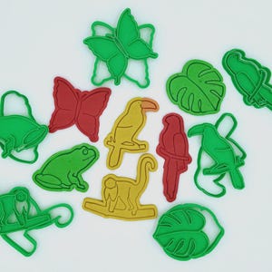 May include: A collection of colorful cookie cutters in various shapes, including a green butterfly, red butterfly, yellow toucan, red parrot, green frog, and a yellow monkey. The cutters are arranged on a white surface.