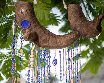 May include: A wooden wind chime with blue and white beads, a blue evil eye charm, and metal chimes. The wind chime is hanging from a tree branch.