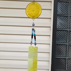 May include: A handcrafted wind chime with a yellow sun-shaped glass disc and two rectangular glass pieces. The chime features a beaded strand with blue, clear, and brown beads. The background is a white siding and a window.