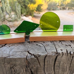 May include: A collection of green glass art pieces in various geometric shapes, including a circle, square, and angled form, displayed on wooden stands. The pieces are translucent and have a textured appearance. The stands are made of light brown wood.