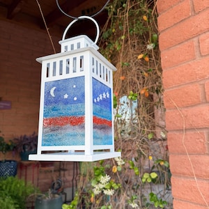 May include: A white lantern with a handle, featuring a painted design of a night sky with a crescent moon and stars, and red and blue stripes. The lantern is hanging outdoors, surrounded by greenery and a brick wall.