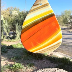 May include: A teardrop-shaped pendant with a vibrant, layered design in shades of orange, yellow, and brown. The pendant is suspended by a silver-colored loop. The background includes a blue sky, an American flag, and desert vegetation.
