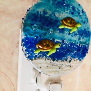 May include: A decorative night light featuring a round design with a blue ocean scene and two sea turtles. The turtles have brown shells and green flippers. The night light plugs into a standard wall outlet.