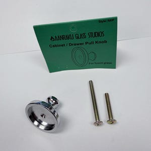 May include: A silver-colored metal cabinet or drawer pull knob for fused glass, accompanied by two metal screws of different lengths. The product is presented with a green display card labeled Aanraku Glass Studios, Style: RKP.
