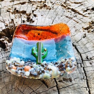 May include: A fused glass dish with a desert theme. The dish features a green cactus in the center, set against a blue and orange background. The base of the dish is filled with small, colorful glass pebbles. The dish is approximately 4 inches wide.