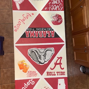 May include: A rectangular folding table painted with University of Alabama themes in crimson and white. Graphics include Bryant-Denny Stadium, a football helmet, an elephant, Nick Saban, the state of Alabama, and text reading Roll Tide.