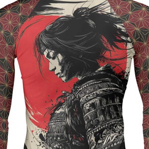 Op de afbeelding: Een compressieshirt met lange mouwen voorzien van een grafische illustratie van een krijger in traditioneel harnas tegen een grote rode cirkel. De mouwen hebben een herhalend geometrisch patroon in zwart, rood en goud, terwijl de romp een zwart-wit profiel toont van een persoon met het haar naar achteren gebonden.