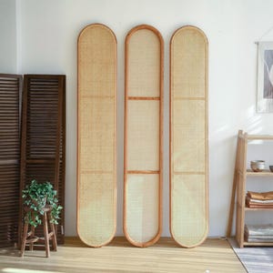 May include: Three tall, arched-top room dividers with woven cane panels and wooden frames. The dividers are a light tan colour and stand against a white wall. A wooden folding screen and a small wooden stool with a plant are visible.