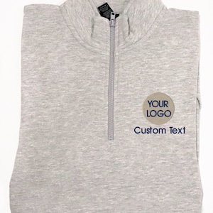 May include: A gray 1/4 zip sweatshirt with a brown circle embroidered with the text "YOUR LOGO" in blue. The text "Custom Text" is also embroidered in blue below the circle.