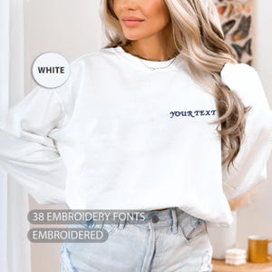 May include: A white long-sleeved sweatshirt with the text "YOUR TEXT" embroidered in navy blue. The image also includes the text "38 EMBROIDERY FONTS" and "EMBROIDERED". The sweatshirt is labeled "WHITE".