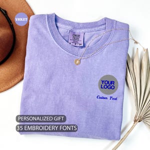 May include: A purple t-shirt with a round gray circle that says "YOUR LOGO" in blue text. Below the circle is the text "Custom Text". The t-shirt is folded and has a tag that says "COMFORT COLORS". The text "PERSONALIZED GIFT" and "35 EMBROIDERY FONTS" is below the t-shirt.