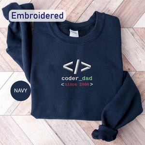 May include: Navy blue sweatshirt with the text "coder_dad <since 2008>" in white and green. The sweatshirt has an embroidered design of code brackets. The word "Embroidered" is in a white rectangle in the upper left corner.