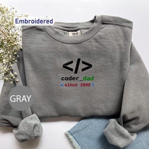 May include: Gray embroidered sweatshirt with the text "coder_dad since 2008" in black, green, red, and blue. The sweatshirt has a crew neck and long sleeves. The word "Embroidered" is above the sweatshirt and the word "GRAY" is below it.