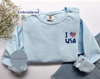 May include: Light blue long-sleeved t-shirt with a red, white, and blue embroidered heart with the text "I USA" on the front. The shirt is made of a soft, chambray fabric.