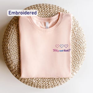 May include: A light pink t-shirt with an embroidered design of three hearts in shades of blue, pink, and purple. The text "Why not both?" is embroidered below the hearts.