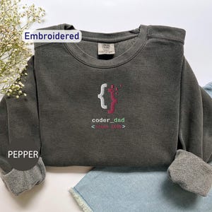 May include: A dark gray sweatshirt with the word "coder_dad" and a graphic of a person inside curly brackets, with the text "since 2006" embroidered on the front. The sweatshirt has cuffed sleeves and the word "Pepper" is printed on the sleeve.