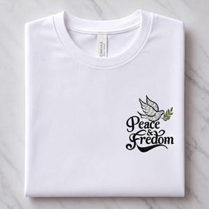 May include: A white short-sleeve t-shirt featuring an embroidered design on the chest. The graphic depicts a grey dove carrying a green olive branch above the black text "Peace & Fredom" written in a stylized, cursive font with a bold flourish.