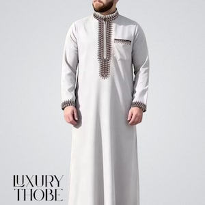 May include: A light gray thobe with intricate brown and black embroidery on the collar, chest, and cuffs. The long-sleeved garment features a small pocket and the text "LUXURY THOBE" at the bottom. The thobe is a traditional Middle Eastern garment.
