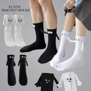 Puede incluir: Calcetines novedosos blancos y negros con diseños de dibujos animados. Presentan brazos y ojos adjuntos, con el texto "FUNNY MAGNET SOCKS". Los calcetines son acanalados y parecen estar hechos de un material suave.