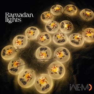 May include: String of Ramadan lights with a warm glow. Each light is a round, frosted glass orb with a crescent moon and lantern design. The words "Ramadan lights" are in the upper left corner.