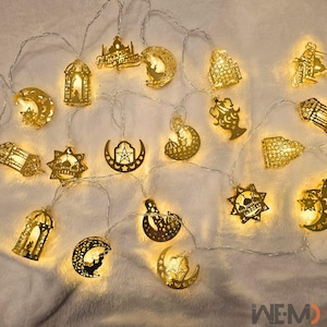 Puede incluir: Una cadena de luces LED blanco cálido con adornos dorados con temática de Ramadán. Los adornos incluyen medias lunas, estrellas y formas de linternas. Las luces están ensartadas en un fino cable plateado, creando una exhibición festiva y decorativa.