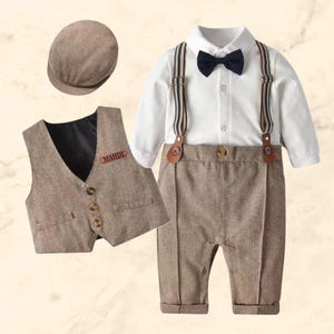 May include: A brown and white baby outfit with a vest, pants, and a bow tie. The vest has a brown leather patch with the name "MAHDE" on it. The outfit also includes a brown newsboy cap.