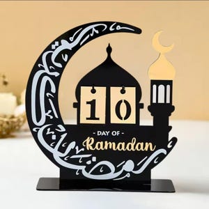 May include: Black metal Ramadan calendar with a crescent moon, mosque, and lantern design. The calendar features a flip-style design with the number 10 displayed. The text "Day of Ramadan" is written below the calendar.