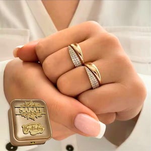 May include: Two gold rings with a curved design, one side encrusted with small, sparkling stones. A gold box with the name "SARAH" and "Happy Birthday" is also visible. The rings are on a hand with manicured nails.