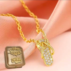 May include: A gold-colored necklace with a flip-flop pendant encrusted with clear gemstones. The necklace is displayed on a pink fabric. A small gold box with the name "SARAH" and "HAPPY Birthday" is also present.